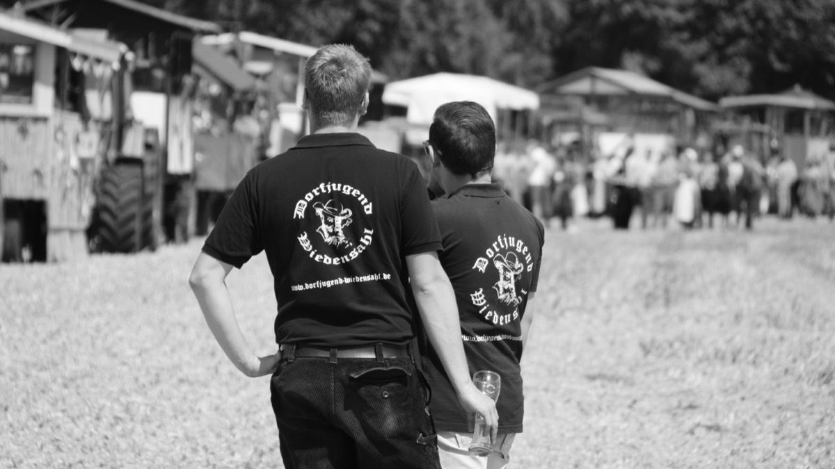 Zwei Mitglieder der Dorfjugend auf dem Festplatz, im Hintergrund Erntewagen und Besucher