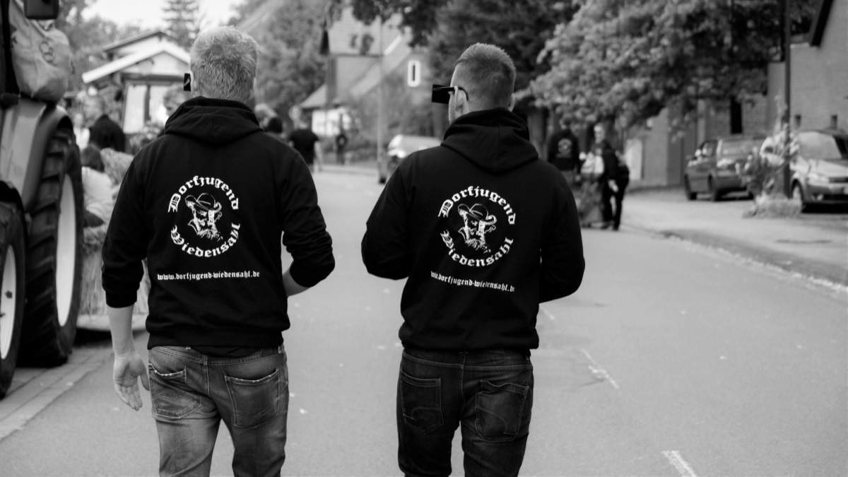Zwei Mitglieder der Dorfjugend im schwarzen Kapuzenpulli mit dem Wappen auf dem Rücken, auf der Dorfstraße beim Erntefest